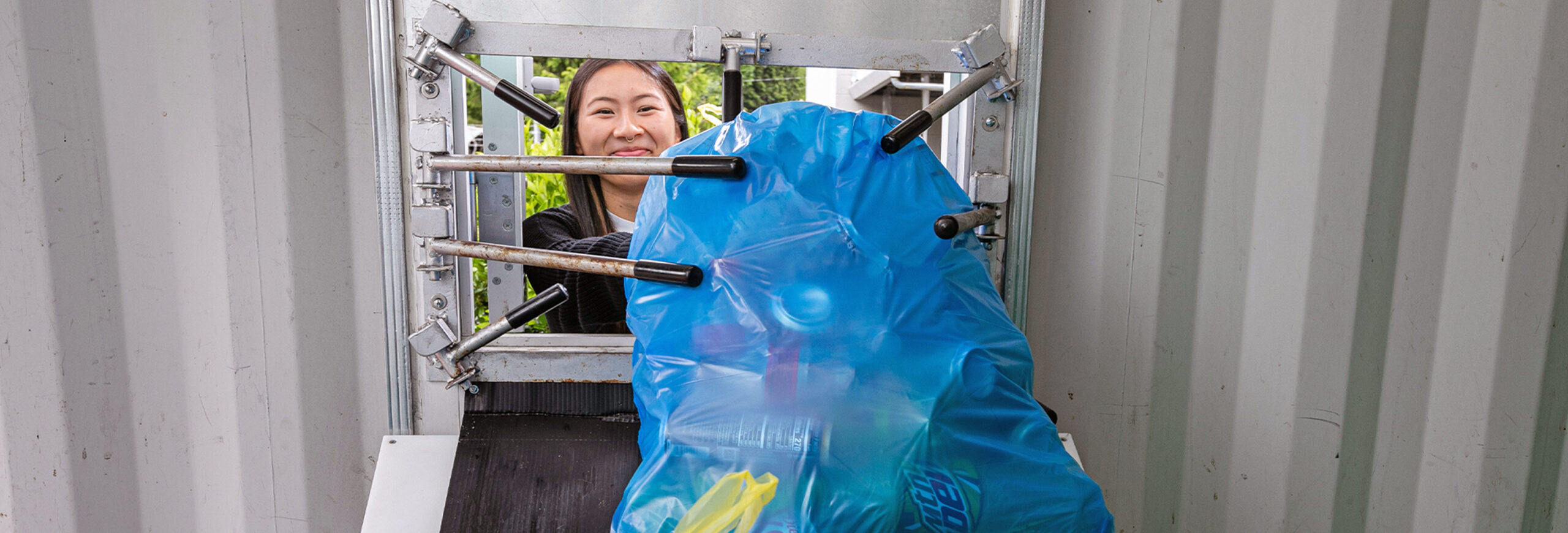 Young woman pushing a blue bag full of recyclable containers into a collection chute.
